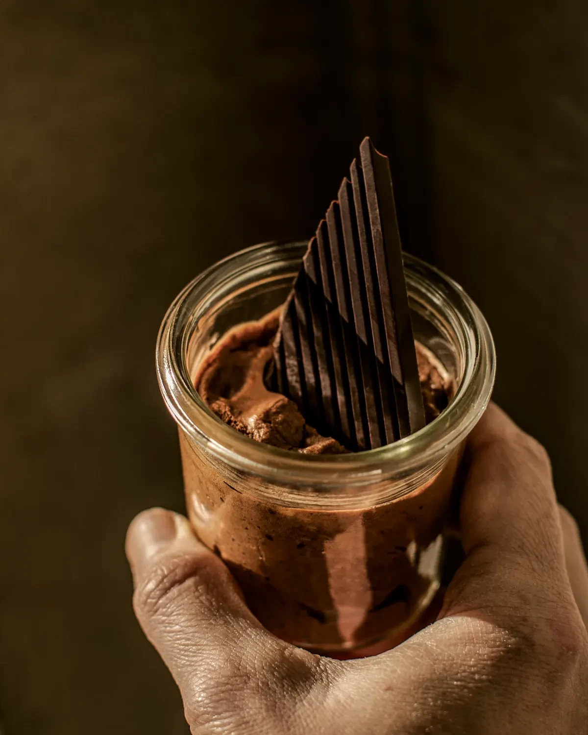 Nahaufnahme einer Hand, die ein Glas mit reichhaltiger Schokoladenmousse hält, garniert mit dünnen, vertikalen dunklen Schokoladensplittern.