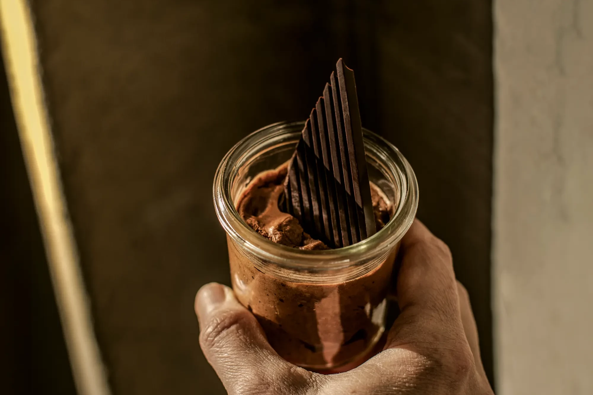 Nahaufnahme einer Hand, die ein Glas mit reichhaltiger Schokoladenmousse hält, garniert mit dünnen, vertikalen dunklen Schokoladensplittern.