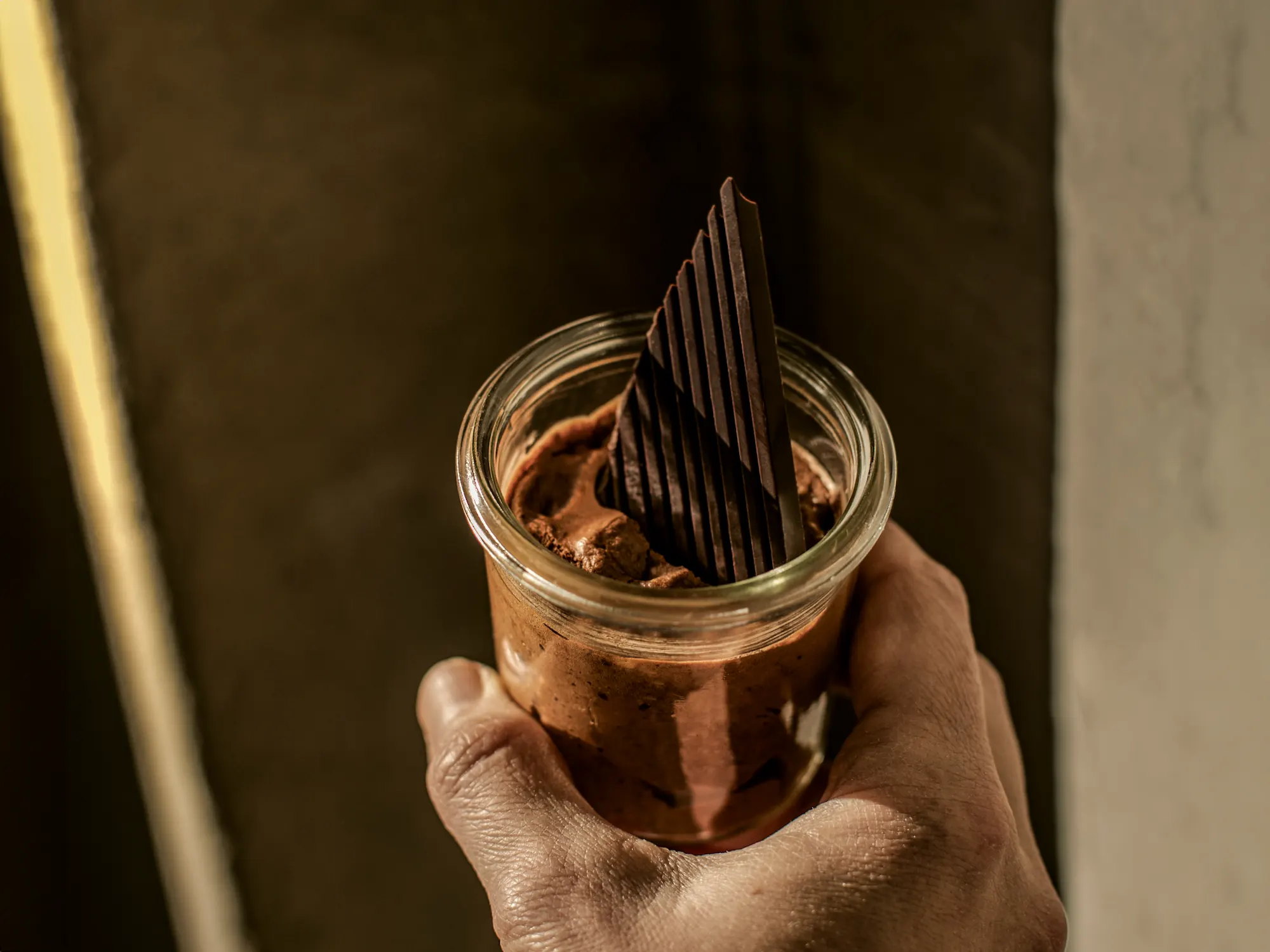 Nahaufnahme einer Hand, die ein Glas mit reichhaltiger Schokoladenmousse hält, garniert mit dünnen, vertikalen dunklen Schokoladensplittern.